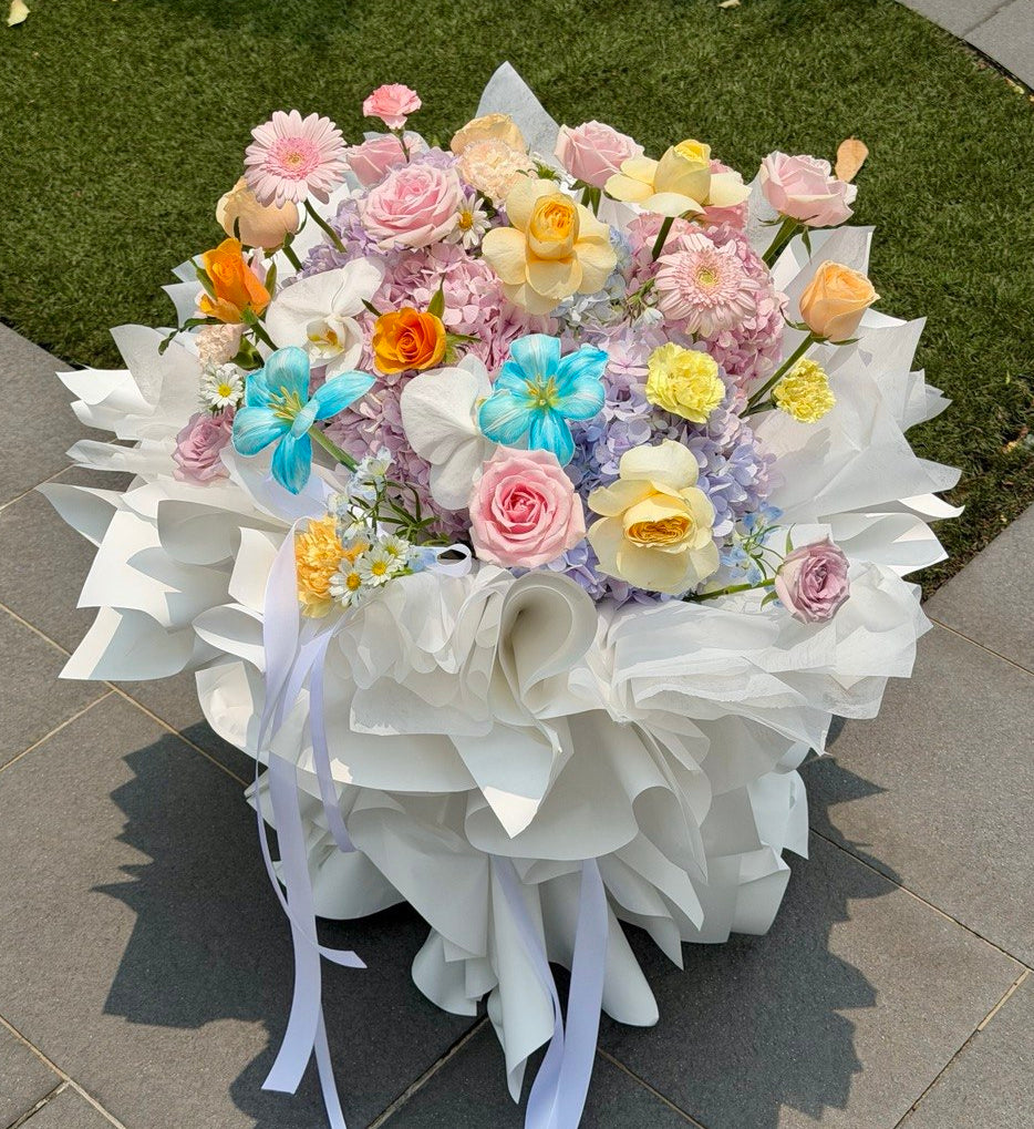 A large, round floral arrangement with a ballet style white paper and white ribbon, filled with pastel pink, yellow, blue, hydrangea and orange flowers, placed outdoors on a paved walkway next to green grass.