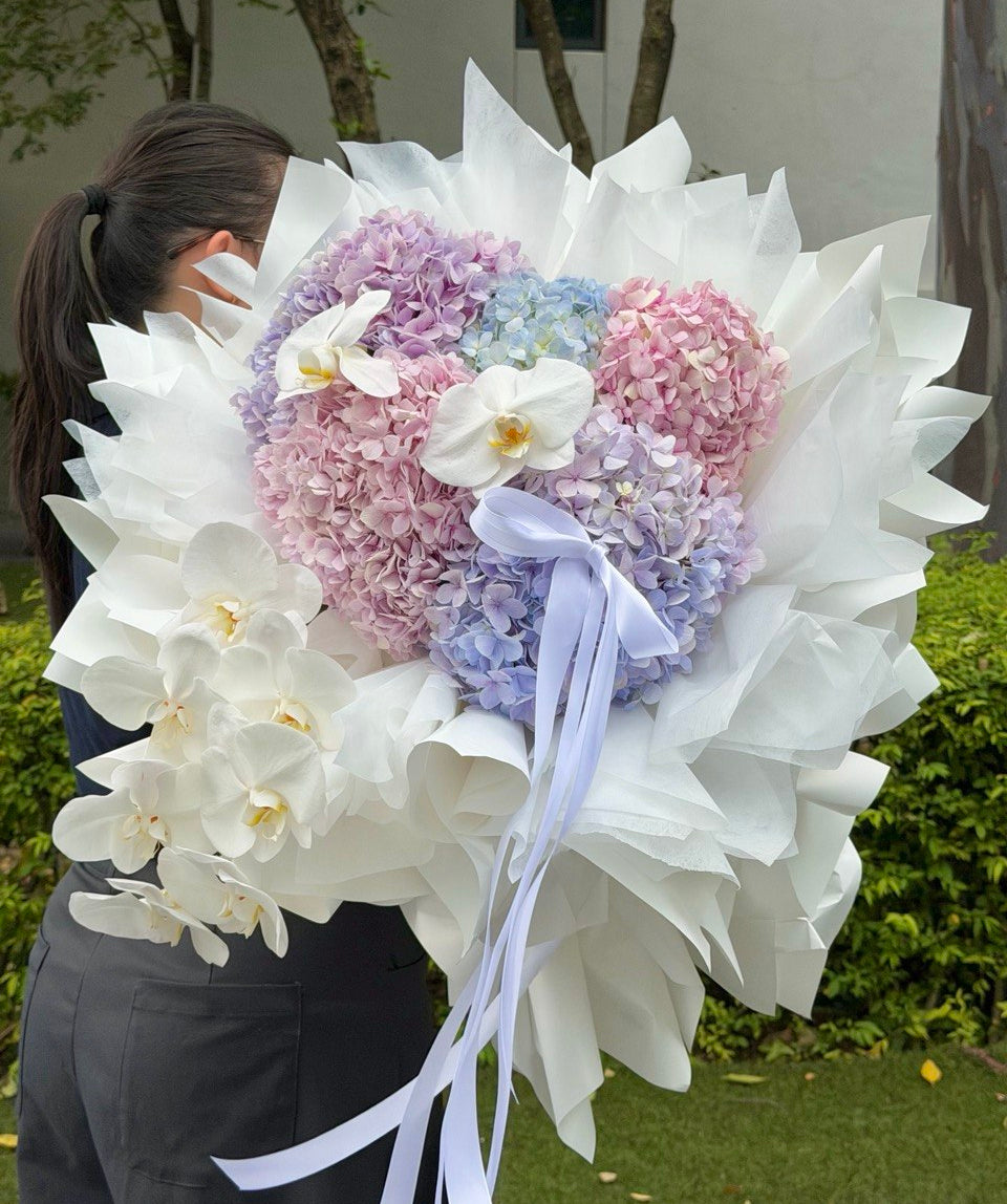 A large heart-shaped bouquet of pastel hydrangeas in pink, purple, and blue, wrapped in white wrapper paper with white orchid flowers and a light blue ribbon, held by a person outdoors.