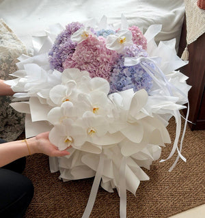 A large floral arrangement featuring white orchids and pastel purple, pink, and blue hydrangeas in the background, wrapped in white wrapper paper with ribbon ties, held by a person on a brown carpeted floor.