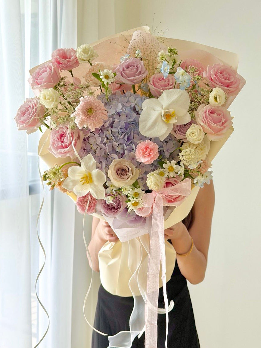 A large, vibrant bouquet of fresh flowers held by a person wearing a black dress. The bouquet features a mix of pink, purple, white, light purple hydrangea and yellow flowers, wrapped in pale pink and cream paper with long white ribbons, set against a bright, softly lit background with sheer white curtains.