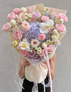 A large, vibrant flower bouquet held by a person against a textured gray wall. The bouquet features a beautiful pastel mix of pink and lilac roses, light purple hydrangeas, white orchids, daisies, and soft pink gerbera daisies, wrapped in elegant cream paper with pale pink ribbons.