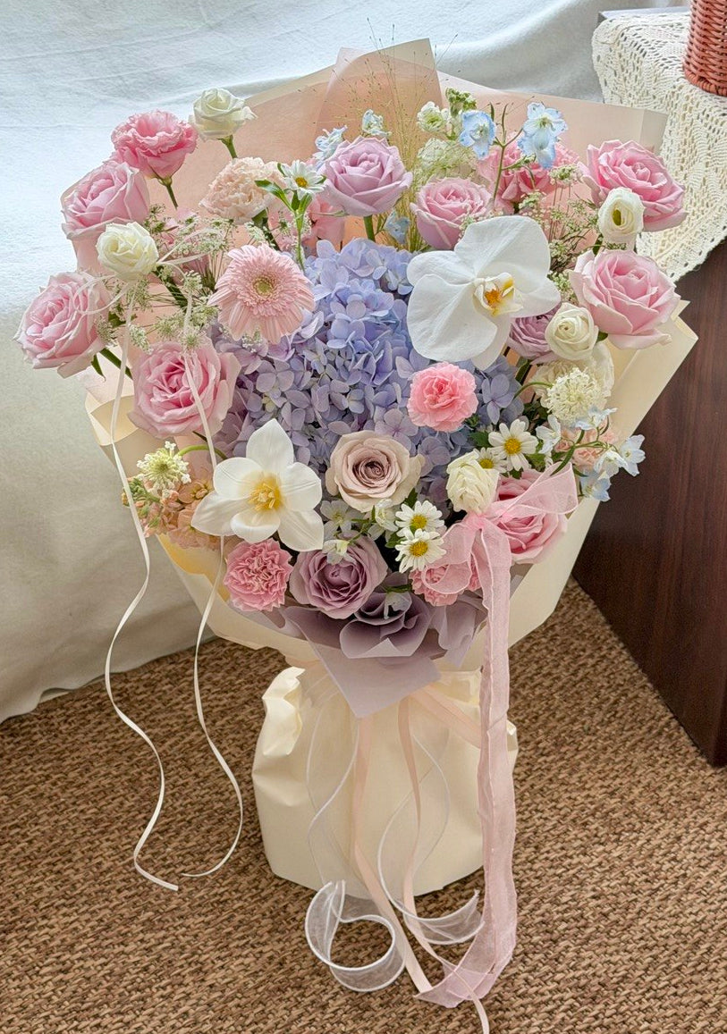 A large, beautifully arranged floral bouquet featuring pink roses, lavender hydrangeas, white orchids, and small white daisies, wrapped in cream paper with pink ribbons, set on a carpeted floor.