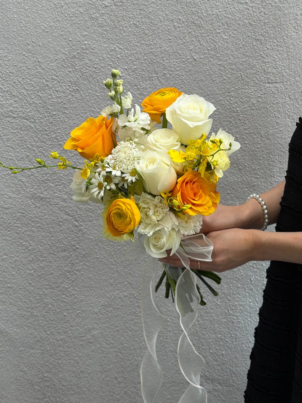 Bouquet of yellow and white wedding flowers held by a person against a gray background for ROM in Malaysia.