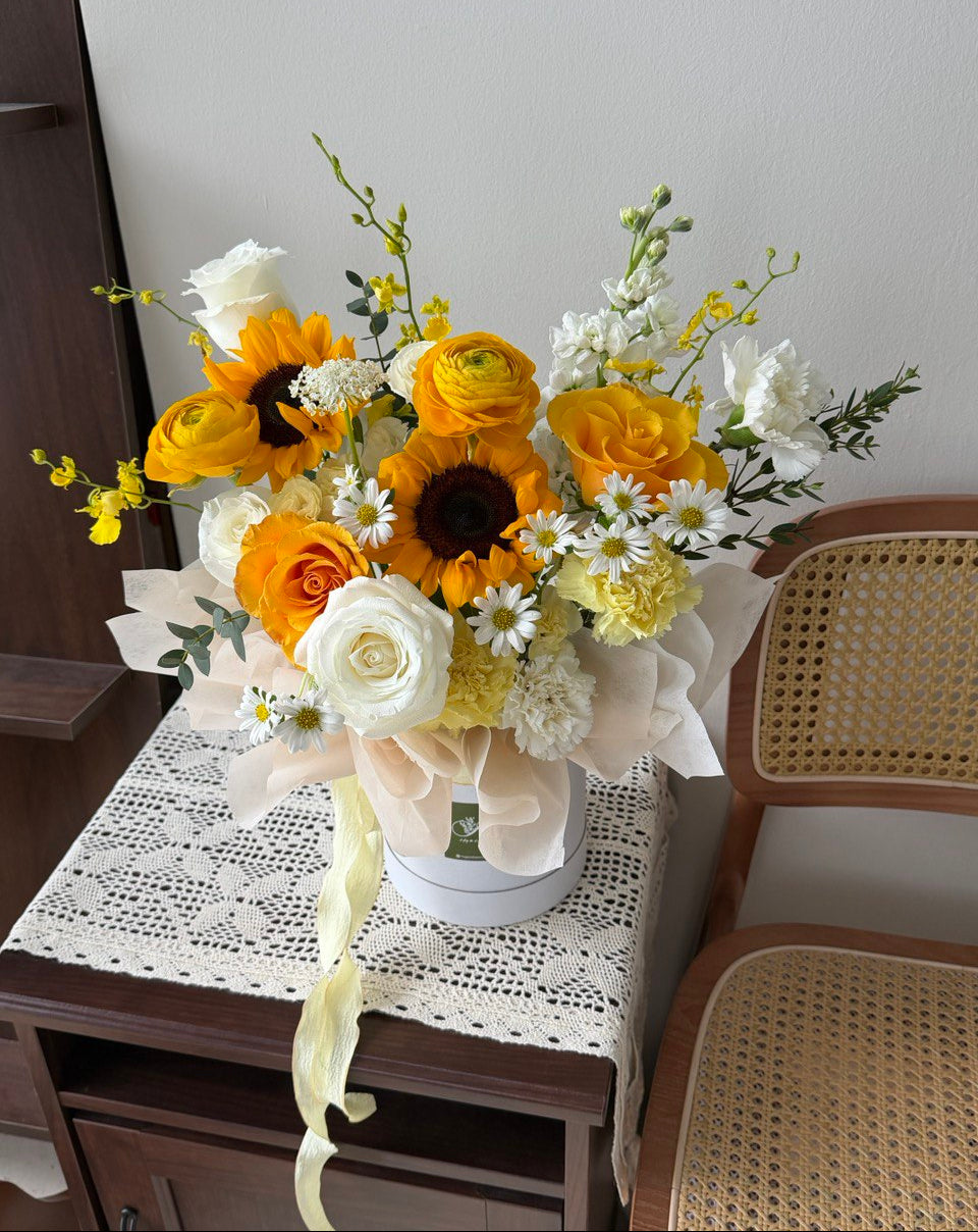 Bouquet of yellow, white, and orange flowers in a box on a table with a chair.