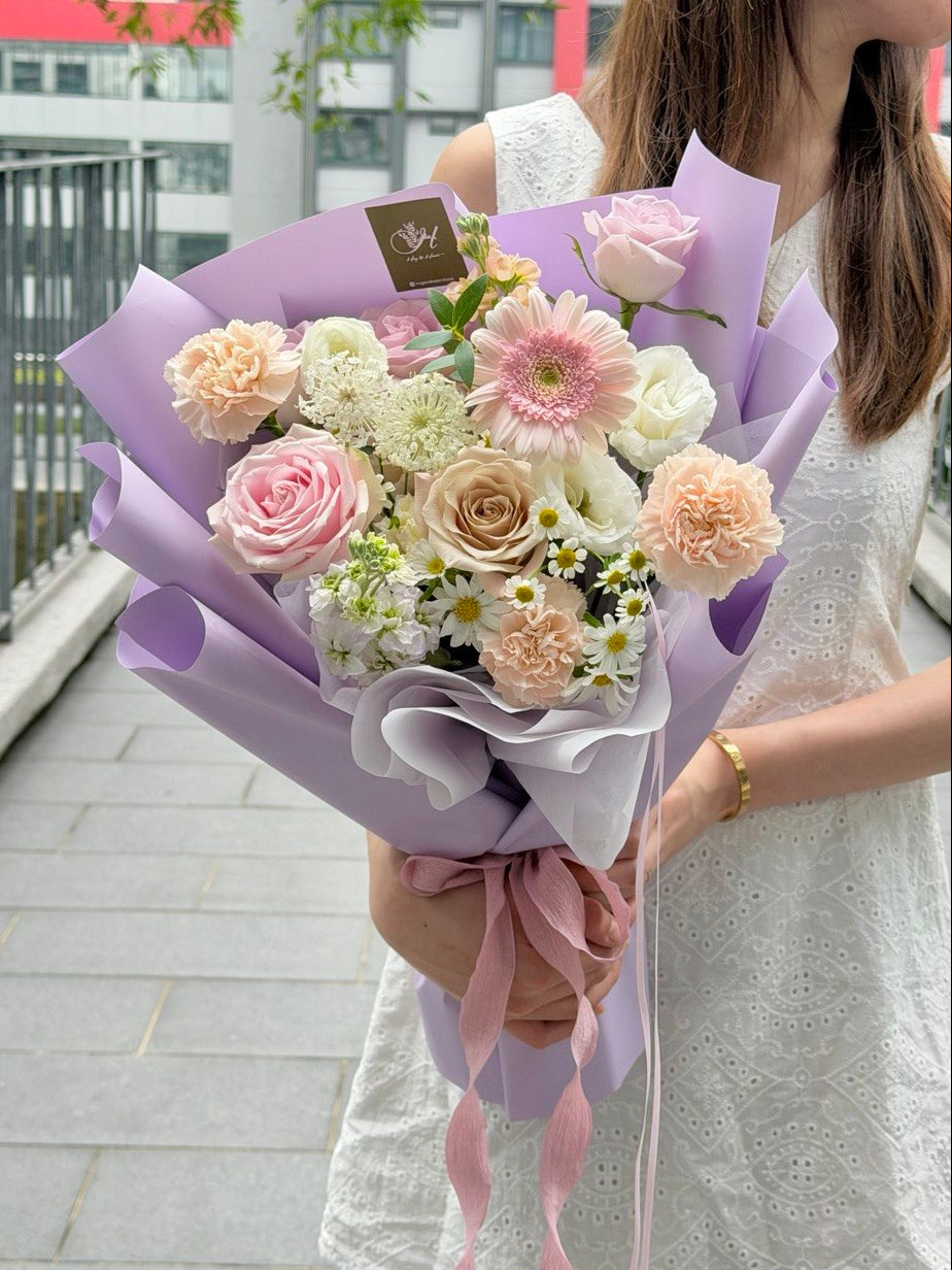 A person in a white dress holds a pastel flower bouquet wrapped in purple paper. The bouquet includes pink, purple, cream, and white flowers with a pink ribbon.