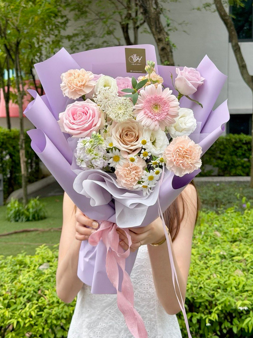 A person outdoors holding a bouquet of pastel flowers wrapped in light purple paper, with greenery and a building in the background.