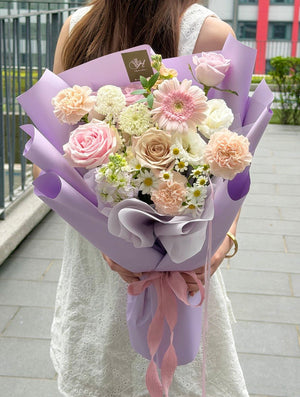 A woman in a white dress holds a bouquet of pink, purple, and white flowers wrapped in light purple paper with a pink ribbon, standing outdoors on a paved walkway.