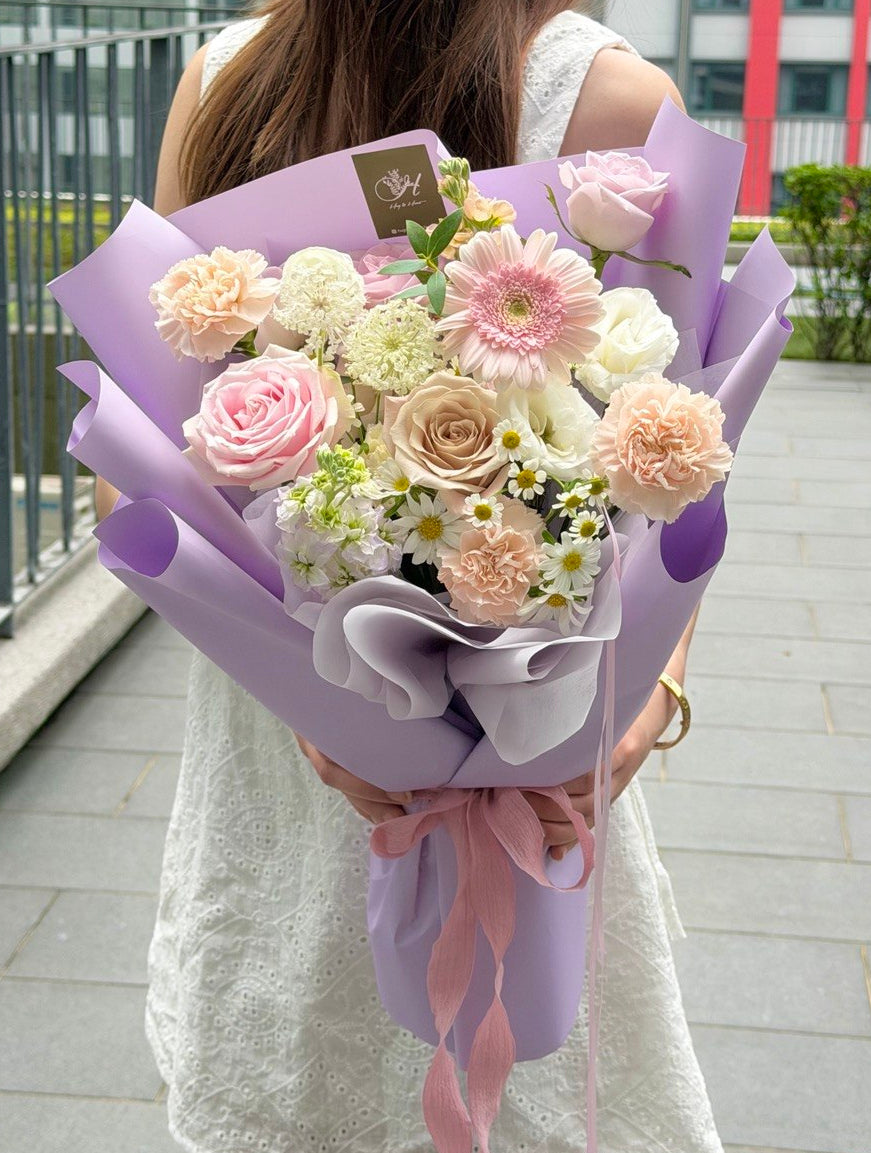 A woman in a white dress holds a bouquet of pink, purple, and white flowers wrapped in light purple paper with a pink ribbon, standing outdoors on a paved walkway.