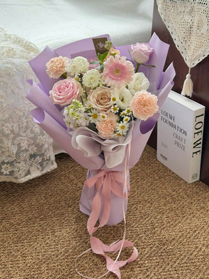 A pastel flower bouquet wrapped in lavender paper and tied with a pink ribbon, placed on a woven rug beside a white lace cushion and a book.