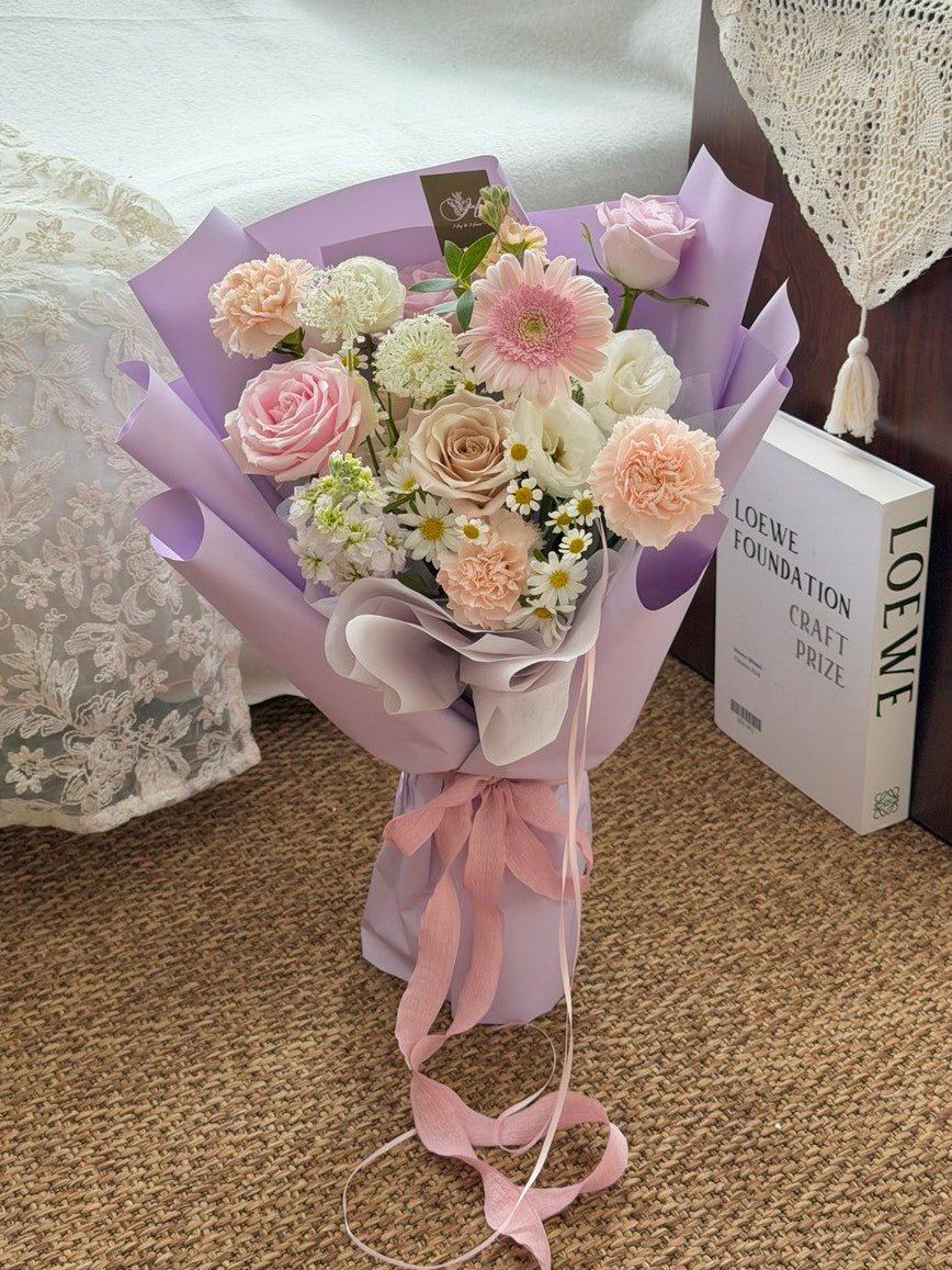A pastel flower bouquet wrapped in lavender paper and tied with a pink ribbon, placed on a woven rug beside a white lace cushion and a book.