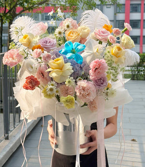 A person outdoors holding a large, vibrant bouquet of multicolored flowers, including pink, yellow, peach, lavender, and blue tulip, placed in a silver flower box with feather accents and ribbons, in a silver bucket vase.