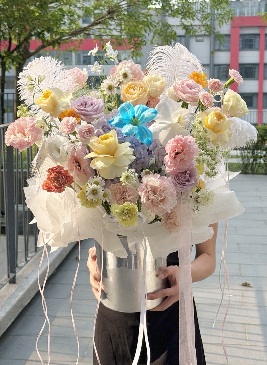 A person outdoors holding a large, vibrant bouquet of multicolored flowers, including pink, yellow, peach, lavender, and blue tulip, placed in a silver flower box with feather accents and ribbons, in a silver bucket vase.