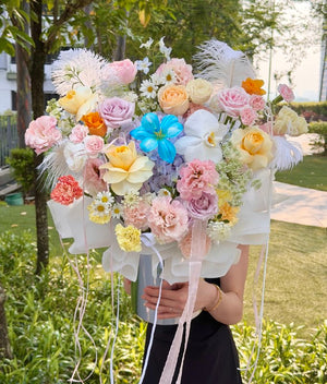 A large, vibrant flower bouquet held by a person in a black dress outdoors. The bouquet features a mix of pastel pink, yellow, orange, and white flowers, including roses, carnations, hydrangeas, and white feathers, wrapped in white tissue paper with long pink ribbons.