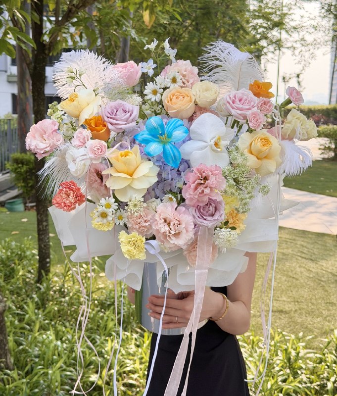 A large, vibrant flower bouquet held by a person in a black dress outdoors. The bouquet features a mix of pastel pink, yellow, orange, and white flowers, including roses, carnations, hydrangeas, and white feathers, wrapped in white tissue paper with long pink ribbons.