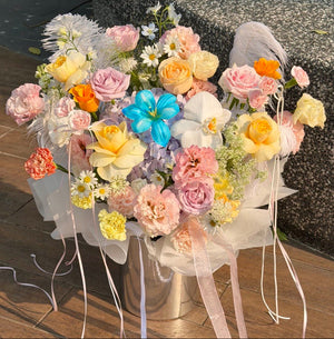 A large, vibrant floral bouquet in a silver metal box, decorated with pastel pink, yellow, peach, and blue flowers, white daisies, and white feathers, tied with long sheer pink and white ribbons, set outdoors on a wooden deck next to a stone bench.