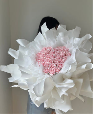 Bouquet of pink soap flower roses wrapped in white paper with a heart shape, held by a person against a neutral background at Subang Jaya with same day delivery.