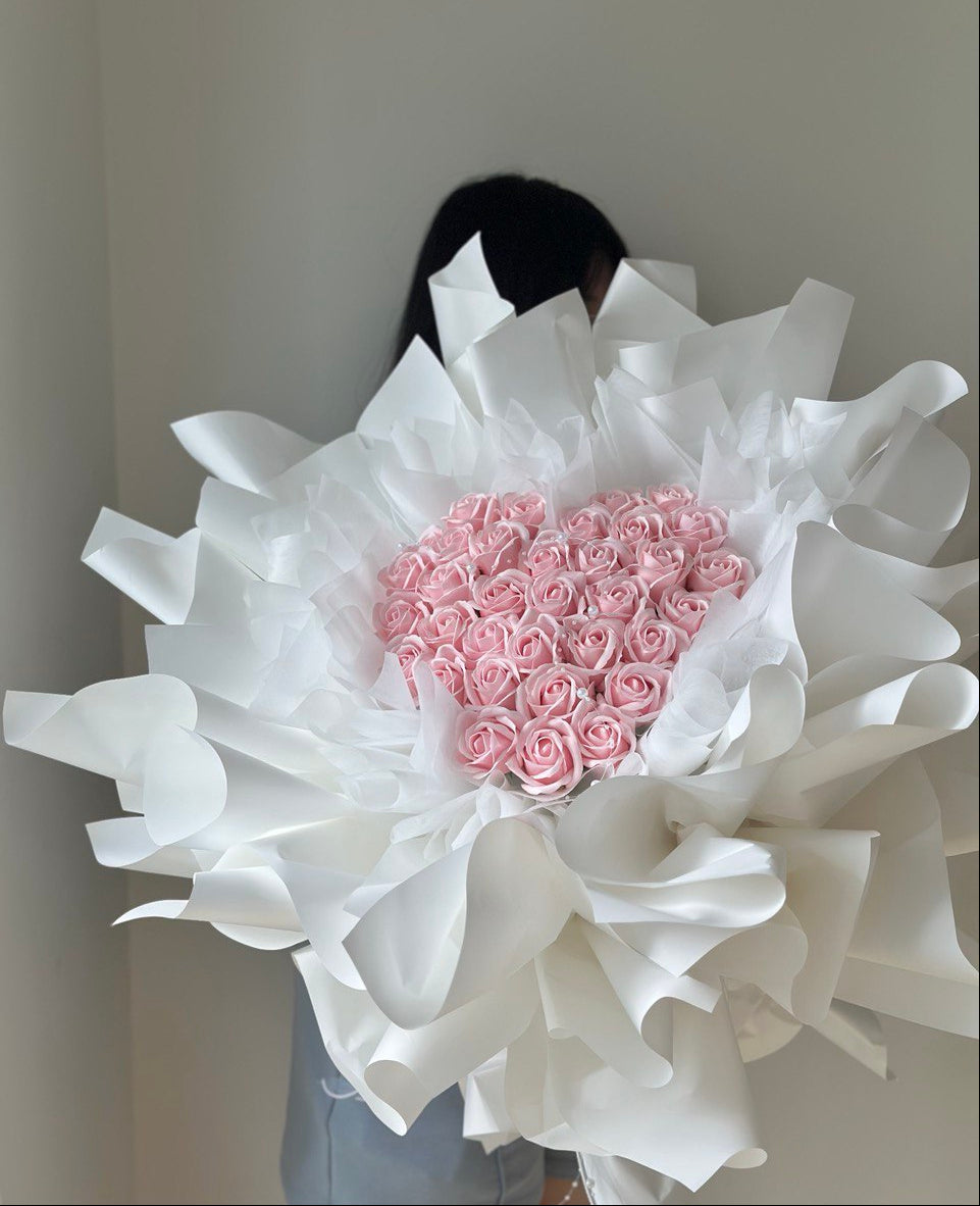 Bouquet of pink soap flower roses wrapped in white paper with a heart shape, held by a person against a neutral background at Subang Jaya with same day delivery.