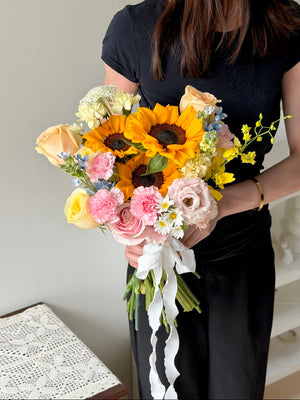 A girl is wearing black holding a bouquet of ROM Sunflower mixed with buttercup rose, champagne rose, pink rose and other fillers with same day delivery at Kuala Lumpur and Selangor.