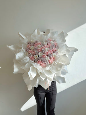 Heart-shaped arrangement of pink and white flowers with white paper flowers on a plain background.