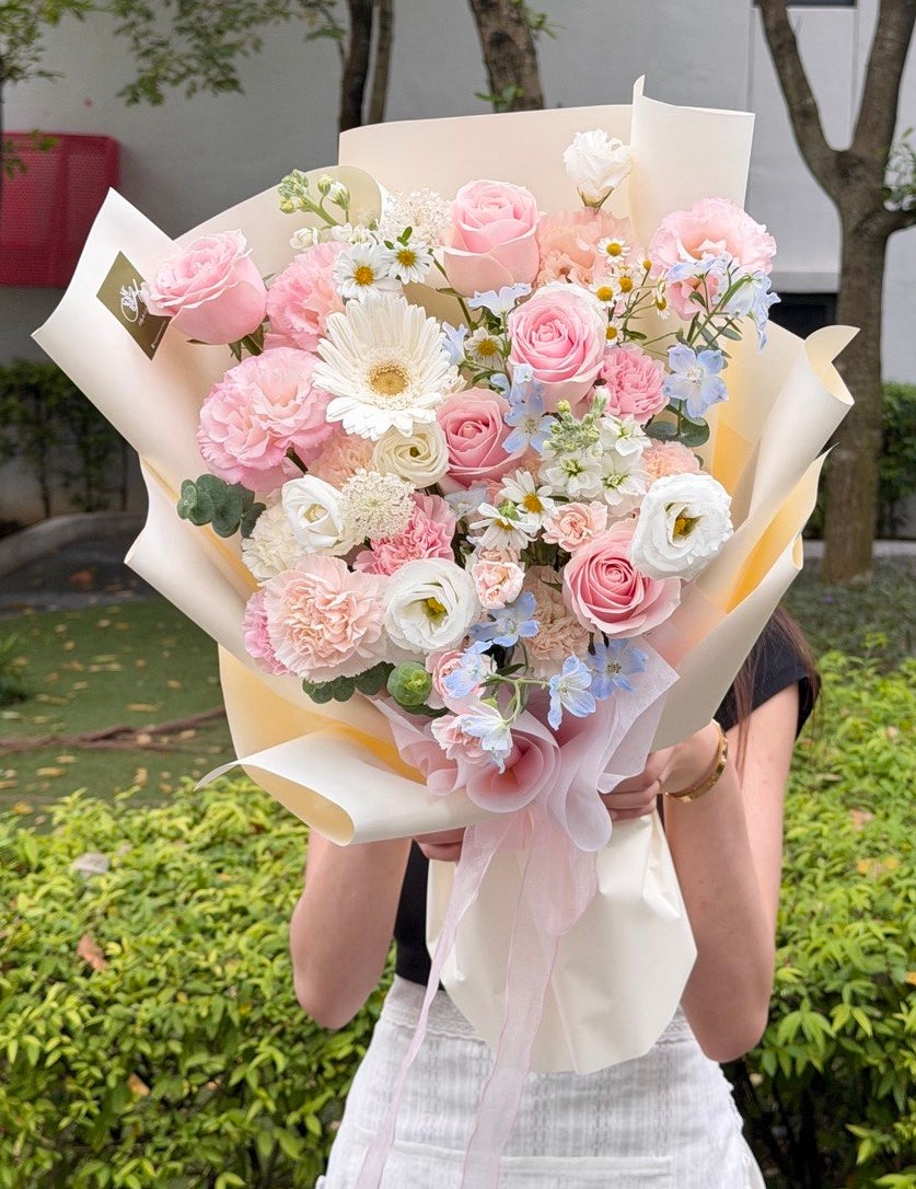 A person outdoors holding a large pastel flower bouquet wrapped in cream paper. The bouquet features pink roses, white daisies, light blue delphiniums, and pink carnations with green foliage.