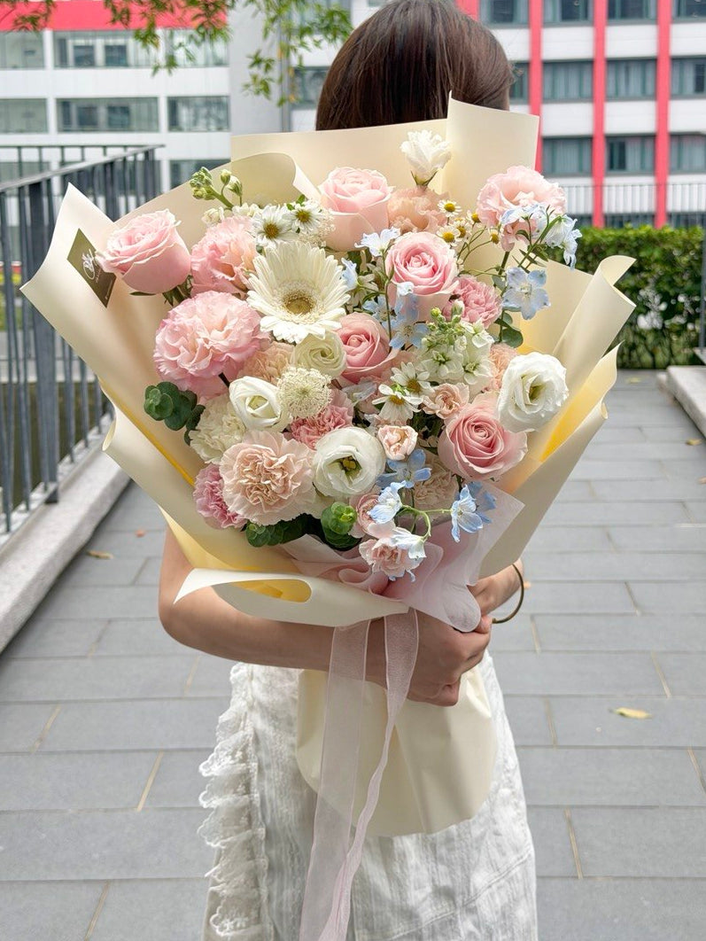 A person standing on an outdoor walkway holding a large bouquet of pink, cream, and light blue flowers wrapped in beige paper. Buildings and trees are visible in the background.
