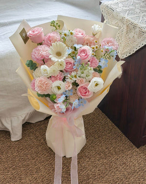 A hand-picked bouquet of pink and white flowers wrapped in cream paper with pink ribbons, placed on a carpeted floor next to a bed.