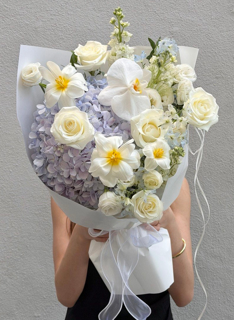 A bouquet of purple hydrangea mixed with tulip, phalaenopsis, white rose held in front of a person wearing a black top against a light gray wall.