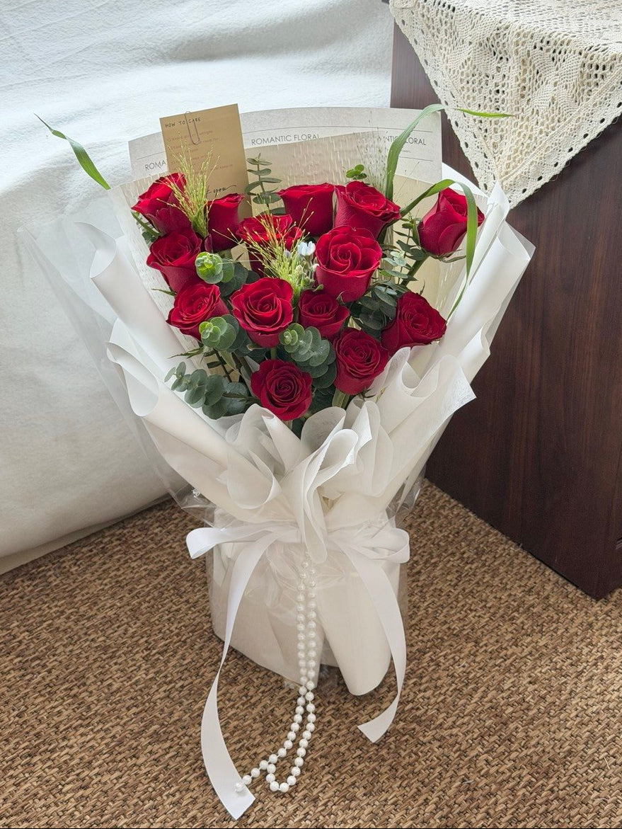 A bouquet of red roses wrapped in white paper and tied with a white ribbon, placed on a carpeted floor.