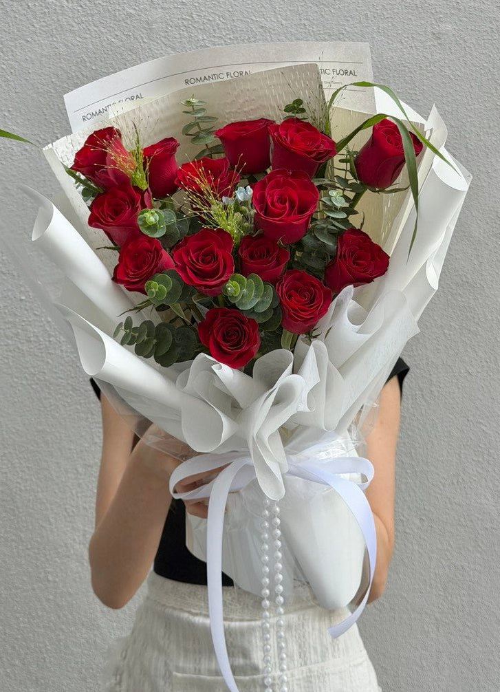 A bouquet of red roses wrapped in white paper with a white ribbon, held by a person whose hands and lower torso are visible against a light gray textured wall.