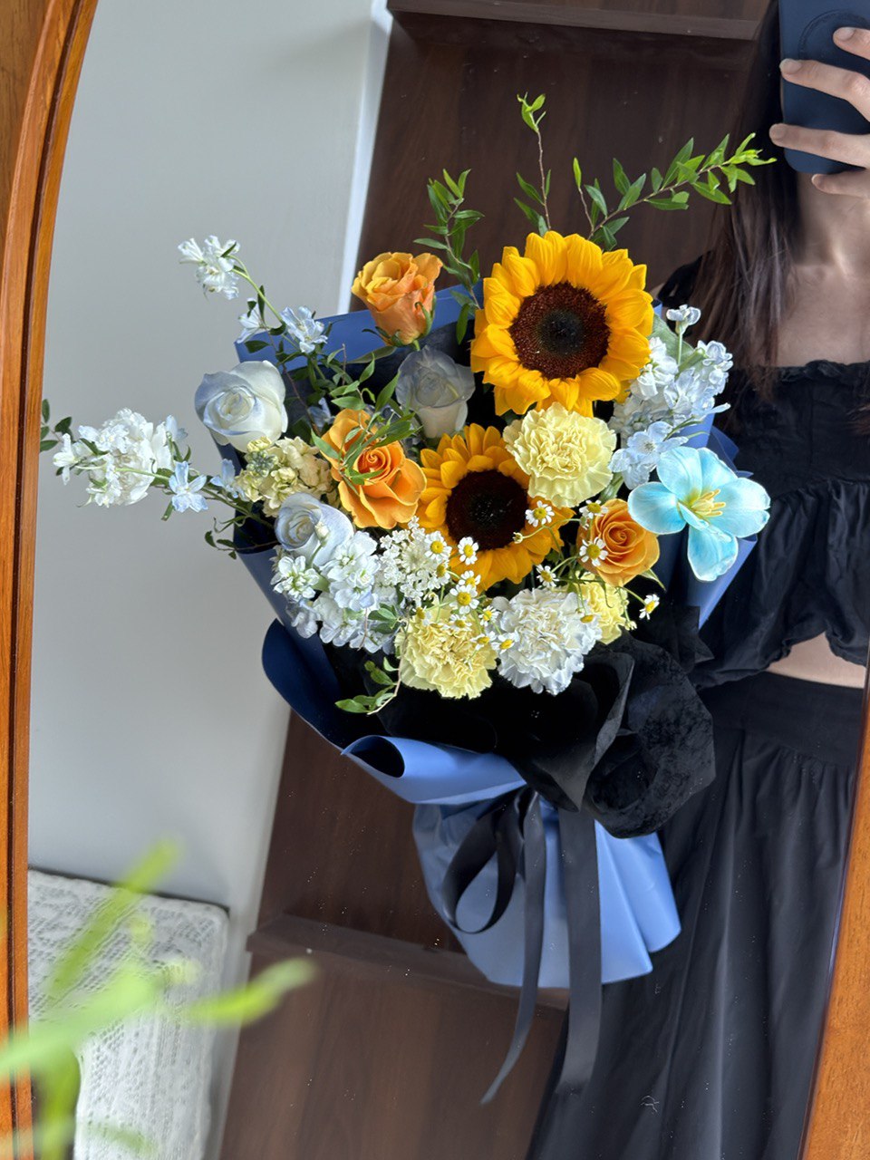 Graduation bouquet with sunflowers, blue roses, yellow carnations and orange roses wrapped in blue paper held by a girl infront of mirror.