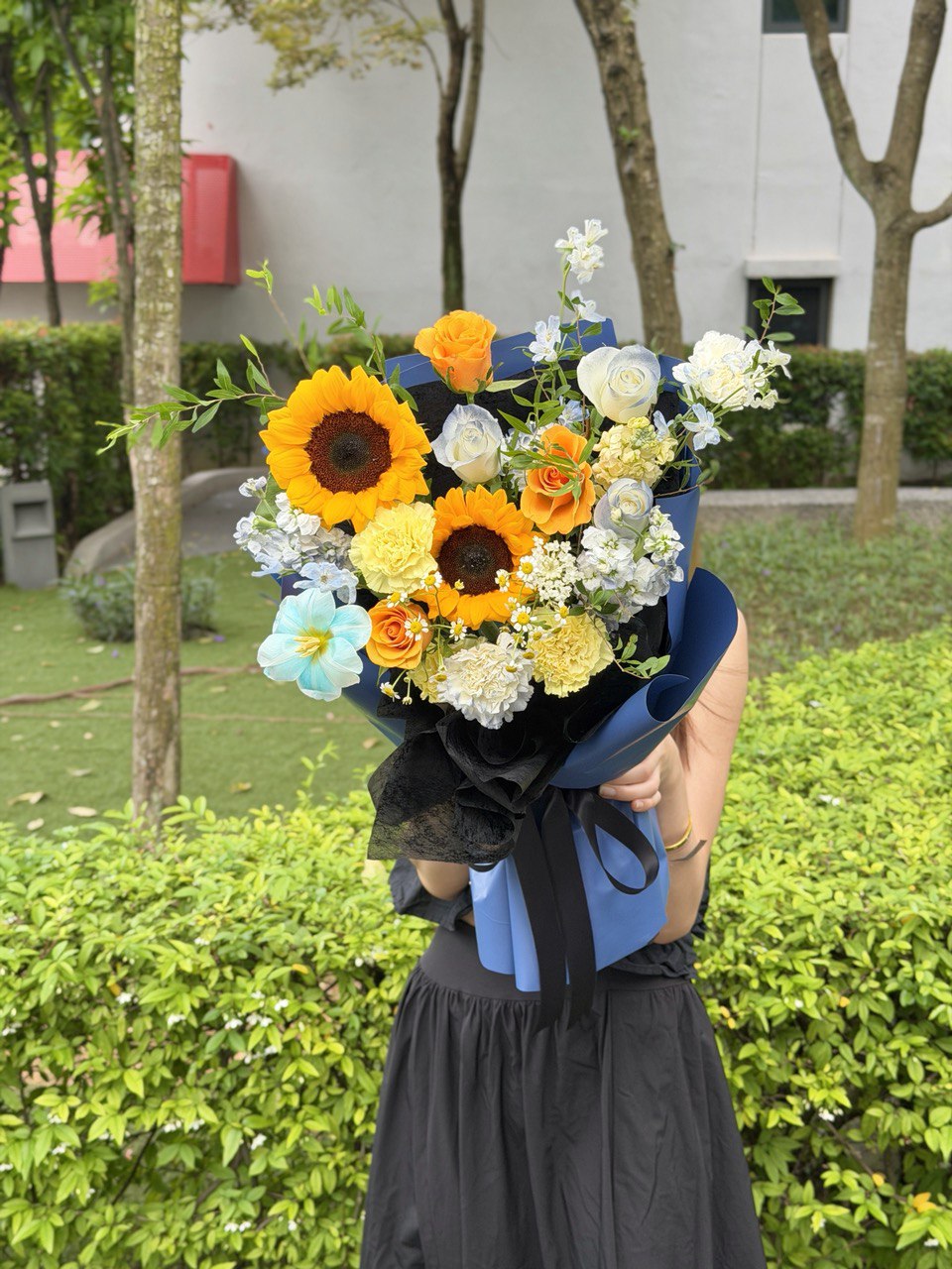Blue and yellow themed graduation flower bouquet featuring sunflowers and roses holding by a girl at outdoor.