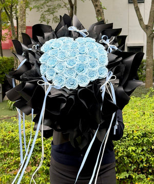 A large bouquet of light blue soap flower roses wrapped in black paper with long white and light blue ribbons, held by a person standing outdoors in front of green bushes and trees.