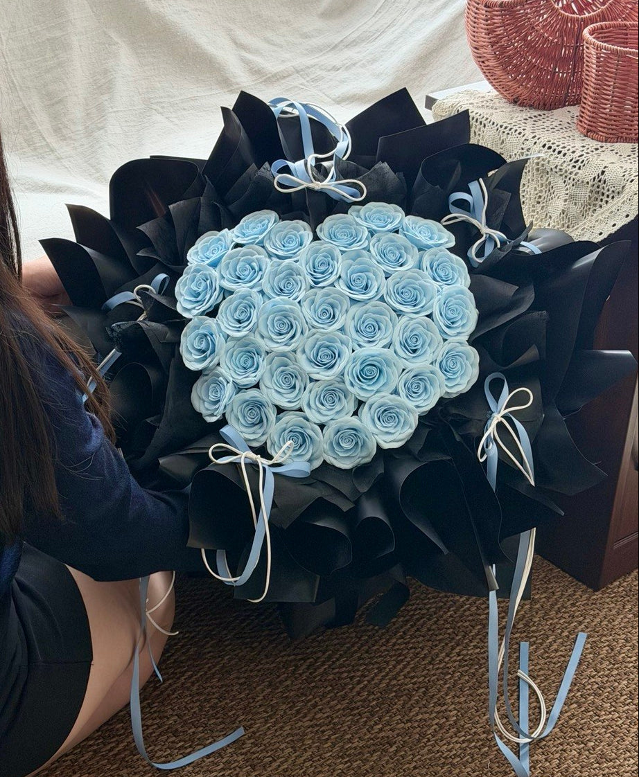 A large bouquet of light blue soap flower roses wrapped in black paper with pale blue ribbons, held by a person seated on a woven floor in an indoor setting.