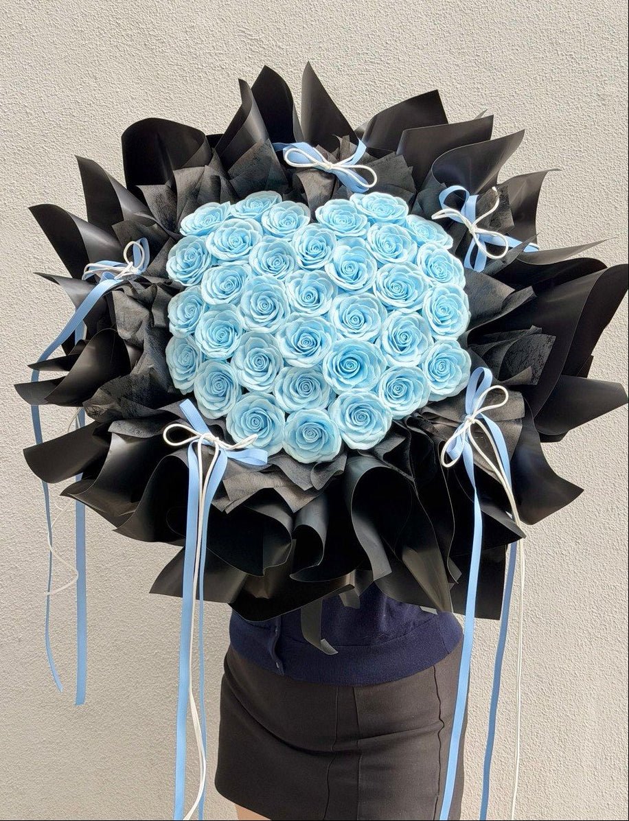 A large bouquet of light blue soap flower roses wrapped in black paper with light blue ribbons, held by a person wearing a navy top and dark skirt against a plain light gray wall.