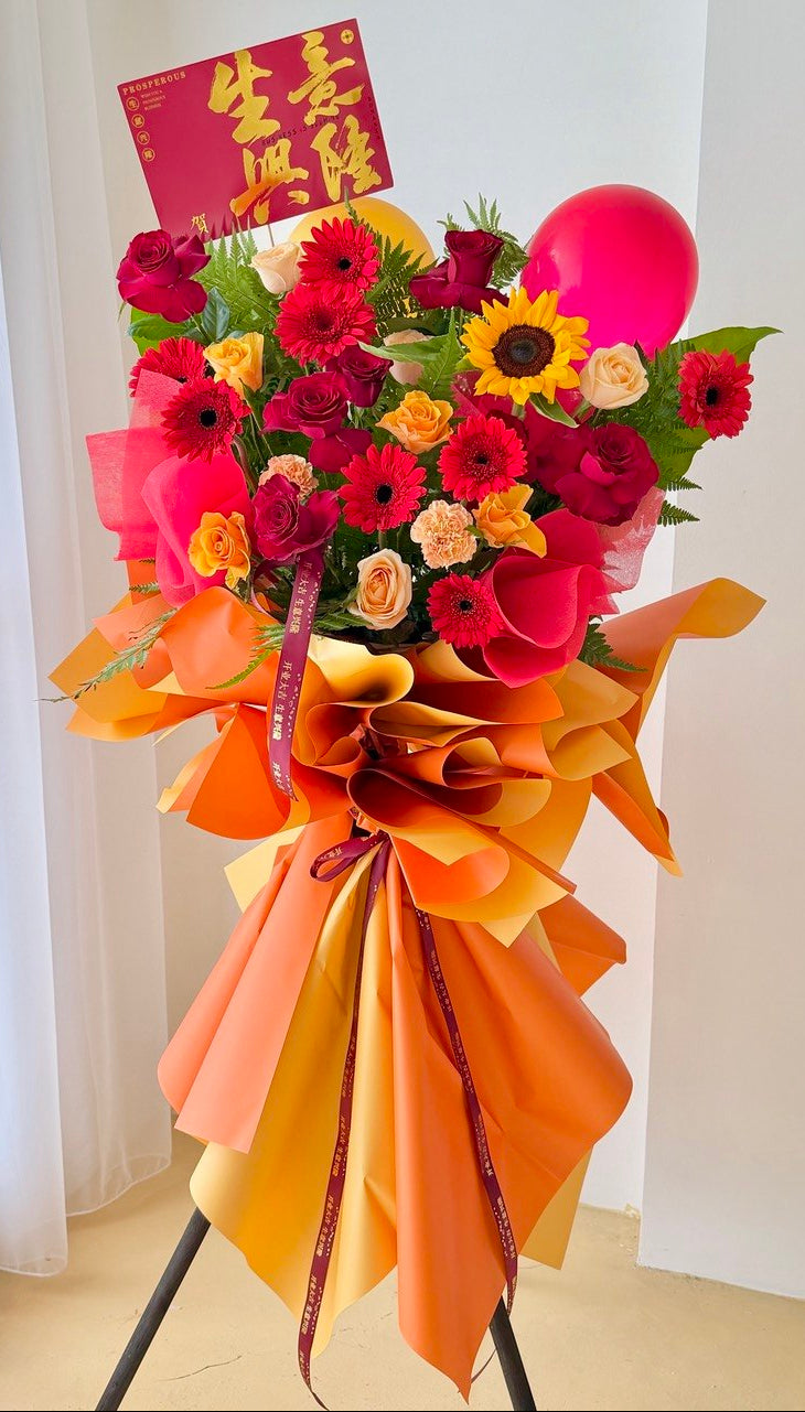 A large celebratory flower arrangement on a stand, featuring red and pink roses, yellow and orange gerbera daisies, a sunflower, a pink balloon, and orange and yellow paper wrapping with a red congratulation card.