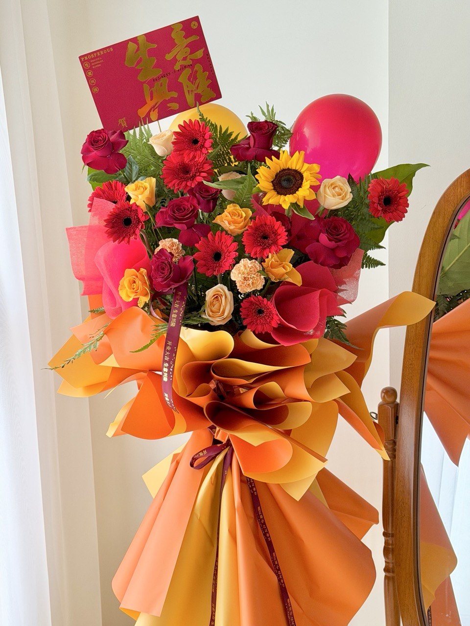 A vibrant business opening flower stand with red, pink, yellow, and orange flowers, a red balloon, and a red Congratulations card, wrapped in orange paper and tied with a ribbon, set next to a wooden mirror.