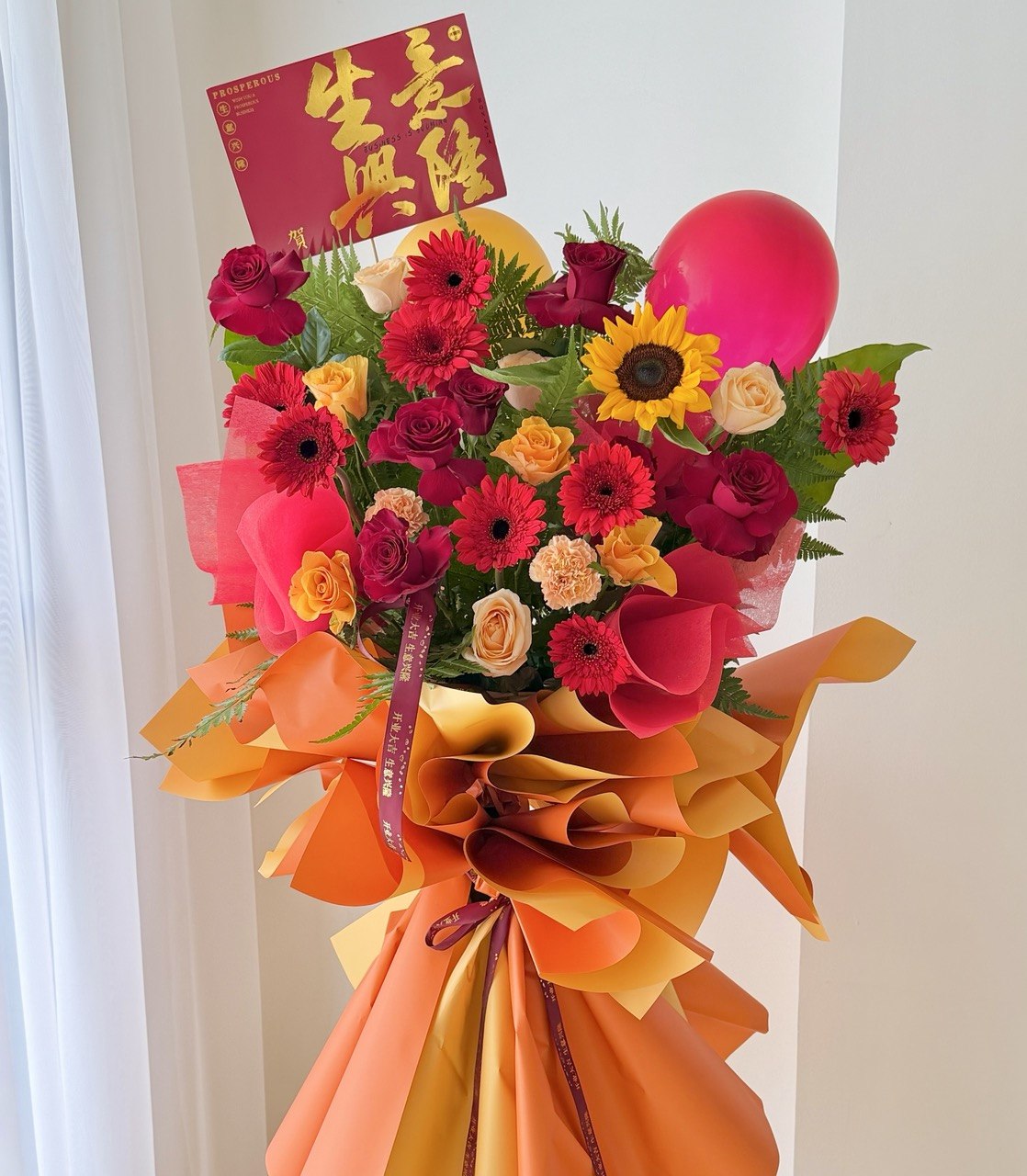 A vibrant business opening flower stand with red, pink, yellow, and orange roses, a large pink balloon, and orange paper wrapping, featuring a red congratulation card.