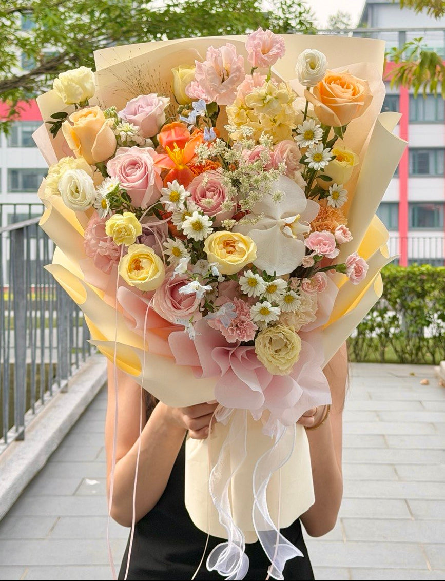 A person holding a large, vibrant flower bouquet with pink, yellow, and white roses, daisies, and orange blossoms, wrapped in cream paper and ribbons, set against an outdoor background of trees and a walkway.