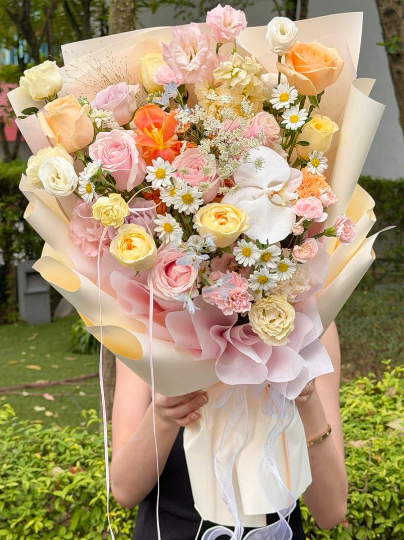 A large, vibrant bouquet of assorted flowers wrapped in cream paper, held by a person outdoors in front of green hedges and trees. The bouquet features a mix of pink, orange, yellow, and white flowers, including roses, daisies, and an orchid.