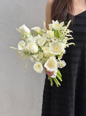 A girl wearing black dress is holding a hand bouquet of calla lily with grey background. 