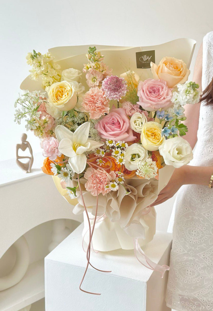 A vibrant bouquet of pastel flowers, including roses, tulip, scabiosa, carnations, chamomile and mixed of other flowers held by a woman in a white embroidered dress.

