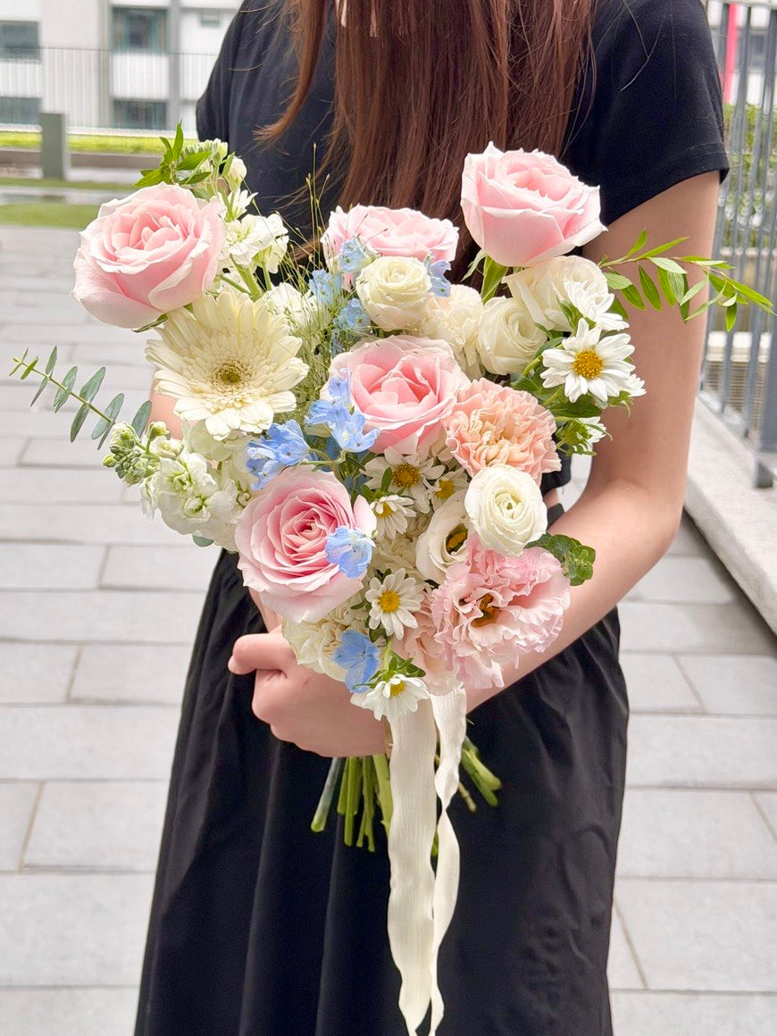 A person in a black dress holds a ROM wedding flower bouquet  featuring pink roses, cream daisies, light blue filler flowers, and green foliage on an outdoor paved walkway.