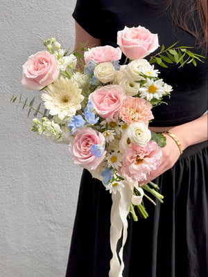 A person in a black dress holds a pastel floral hand wedding bouquet featuring pink roses, cream daisies, light blue flowers, and green foliage against a light gray wall.