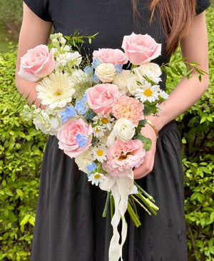 A person in a black dress holds a bouquet of wedding flower featuring pink, white, and light blue flowers outdoors in front of green bushes.