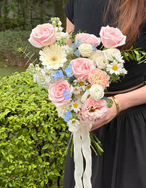 A person in a black dress holds a pastel floral bouquet featuring pink and white roses, white daisies, light blue ranunculus, and green foliage, with long white ribbon ties, set against a green hedge background.