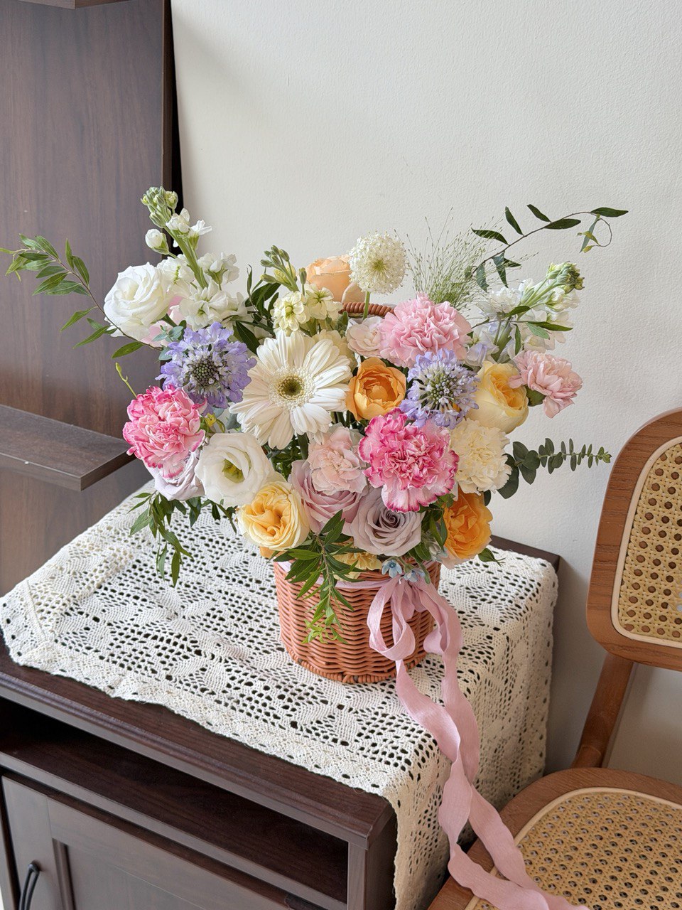 Bouquet of flowers in a basket on a lace tablecloth with a chair in the background.