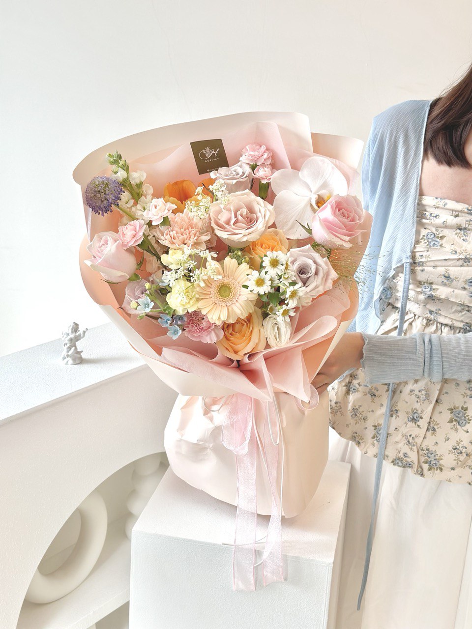 Close-up of a pastel-toned flower bouquet with roses, gerberas, and mixed flowers, held by a woman dressed in a light blue cardigan and floral outfit.