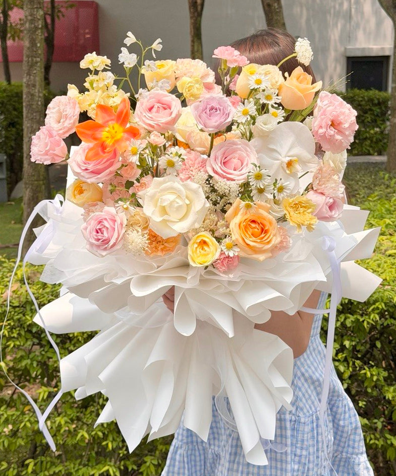 A large, vibrant floral bouquet with pink, peach, yellow, and white flowers wrapped in white paper, held by a person wearing a blue and white gingham dress in an outdoor garden setting.