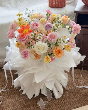 A large, vibrant floral arrangement of pastel pink, peach, yellow, and purple roses with white daisies, wrapped in a voluminous white paper wrapper with long ribbons, set on a brown carpeted floor.