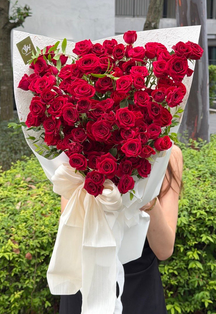 A large bouquet of deep red rose rose spray wrapped in white paper with a white ribbon bow, held in front of a person outdoors in front of green bushes.